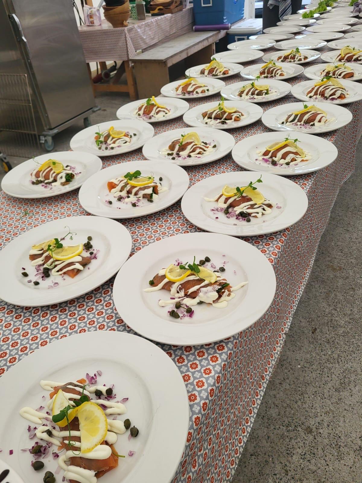 Dozens of smoked salmon starters plated in rows on a long table, each with dill creme fraiche, capers, diced red onion, a lemon slice and a herb sprig, ready to be carried in