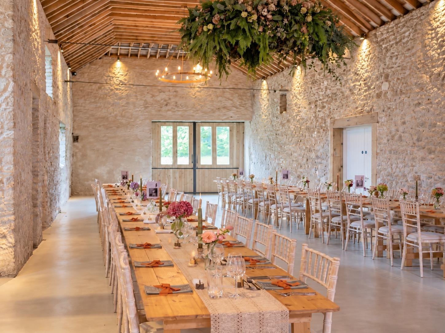 Juniper Barn set for the wedding feast: long timber tables with runners, flowers and candles under the stone walls and hanging greenery