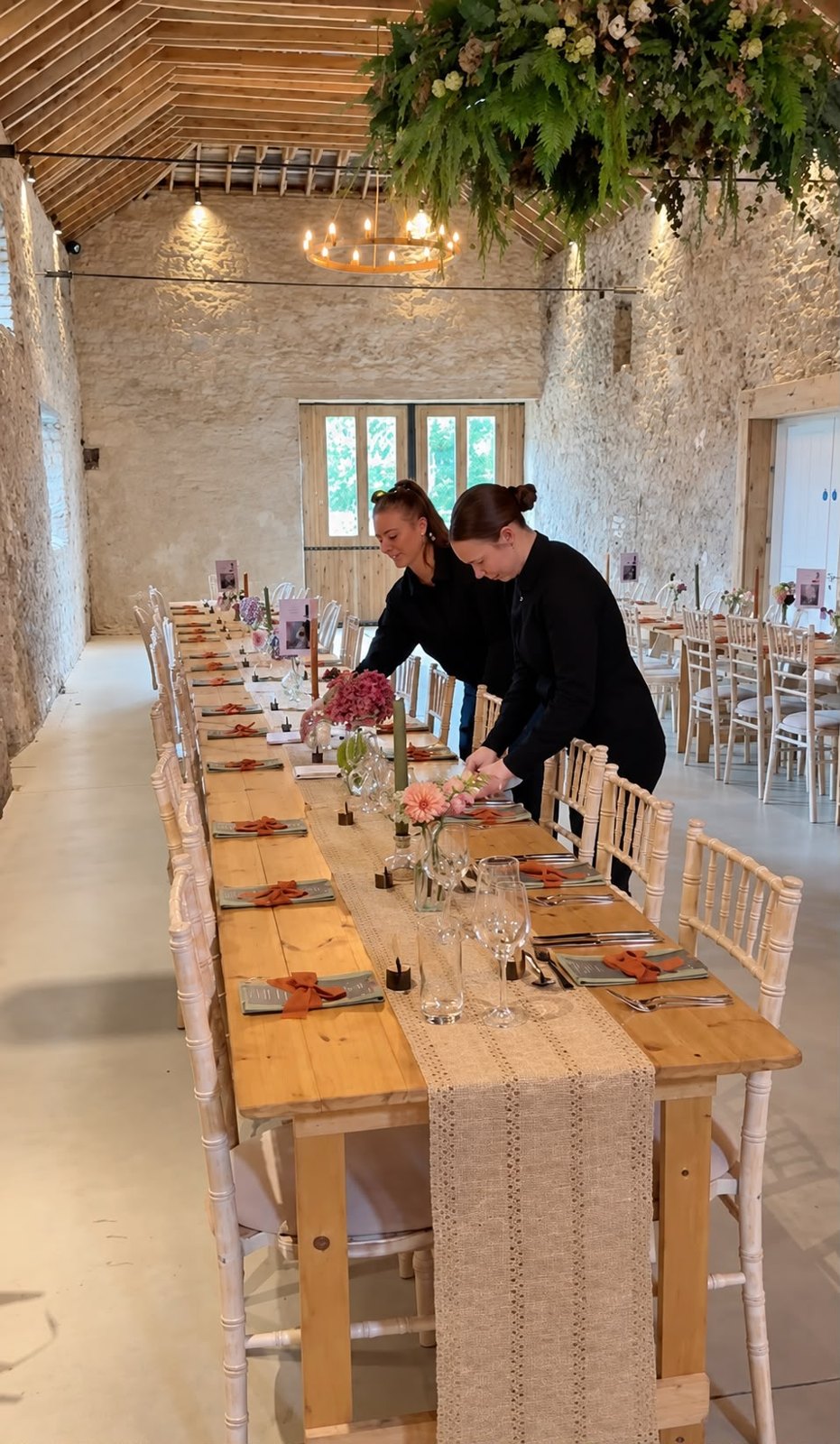 Two of the Honestly team laying the long table at Juniper Barn, with the ring chandelier and hanging greenery above