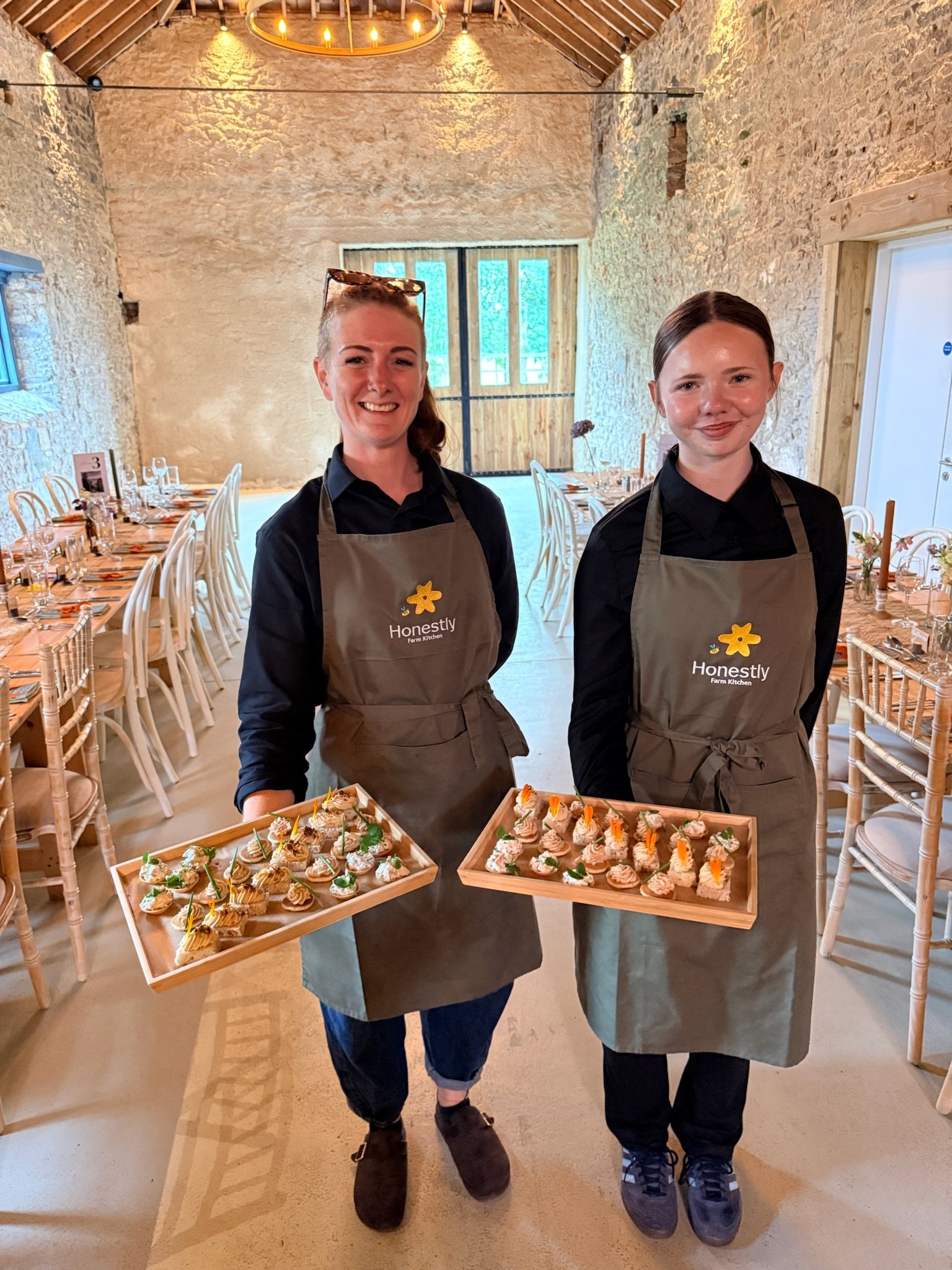Two of the Honestly team in the barn with trays of canapés, the long tables set behind them