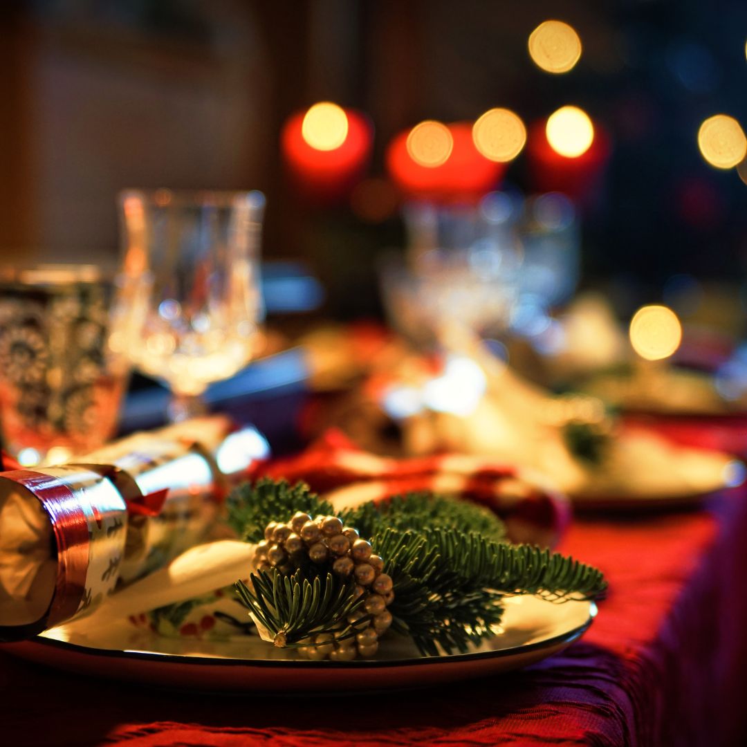 A festive place setting with a Christmas cracker, a sprig of pine and a gold pine cone on the plate, candles glowing behind